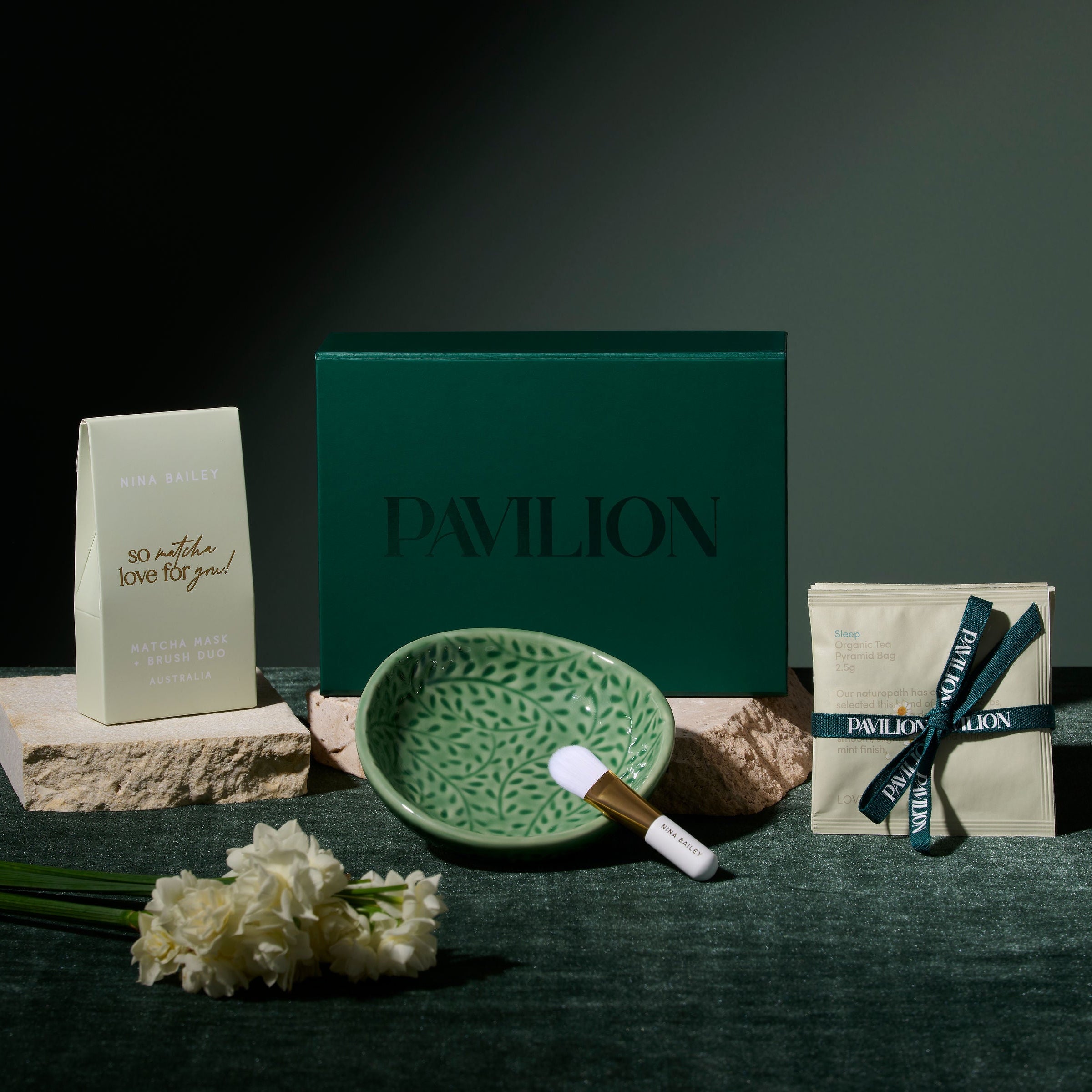 Pavilion skincare gift set arranged on a dark green velvet surface against a dark background. The set includes a large dark green box with the word 'PAVILION' , a matcha clay mask, a small green patterned ceramic bowl, a white and gold mask brush, and a trio of beauty teas.