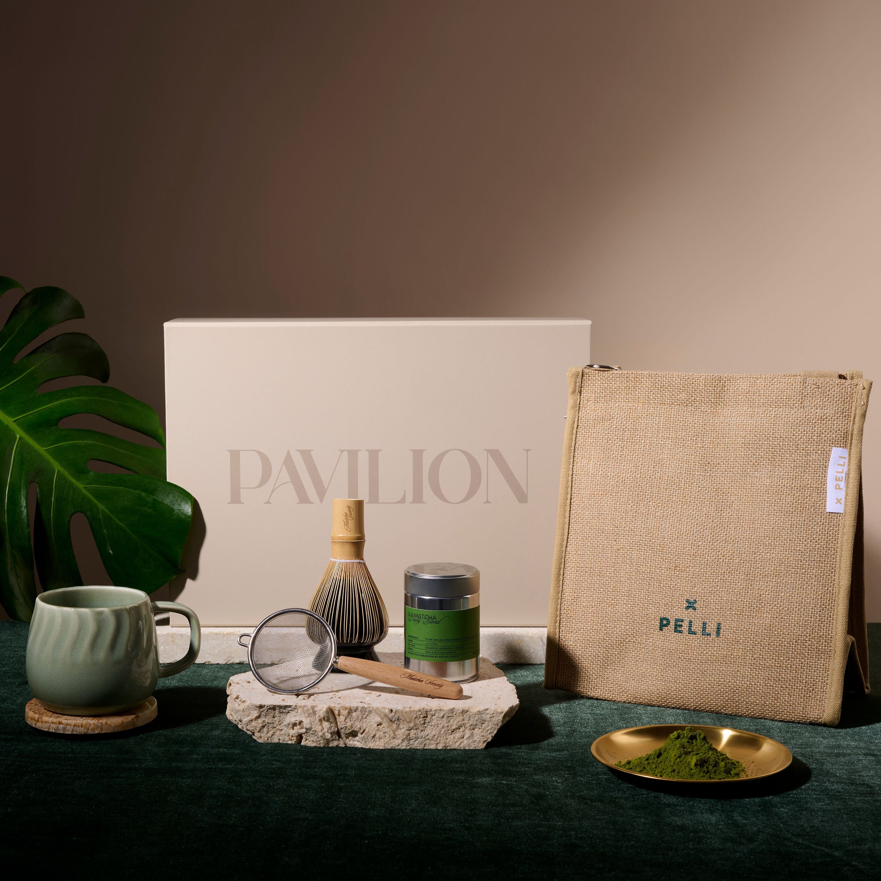 A styled tea gift set on a dark green velvet surface against a warm beige background. The set includes a large beige box with 'PAVILION' printed on it, a beige jute pouch with 'PELLI' printed on it, a green ceramic mug, a bamboo whisk, a small glass scoop, a jar of green tea, and a small gold dish of matcha powder, accompanied by a large green monstera leaf.