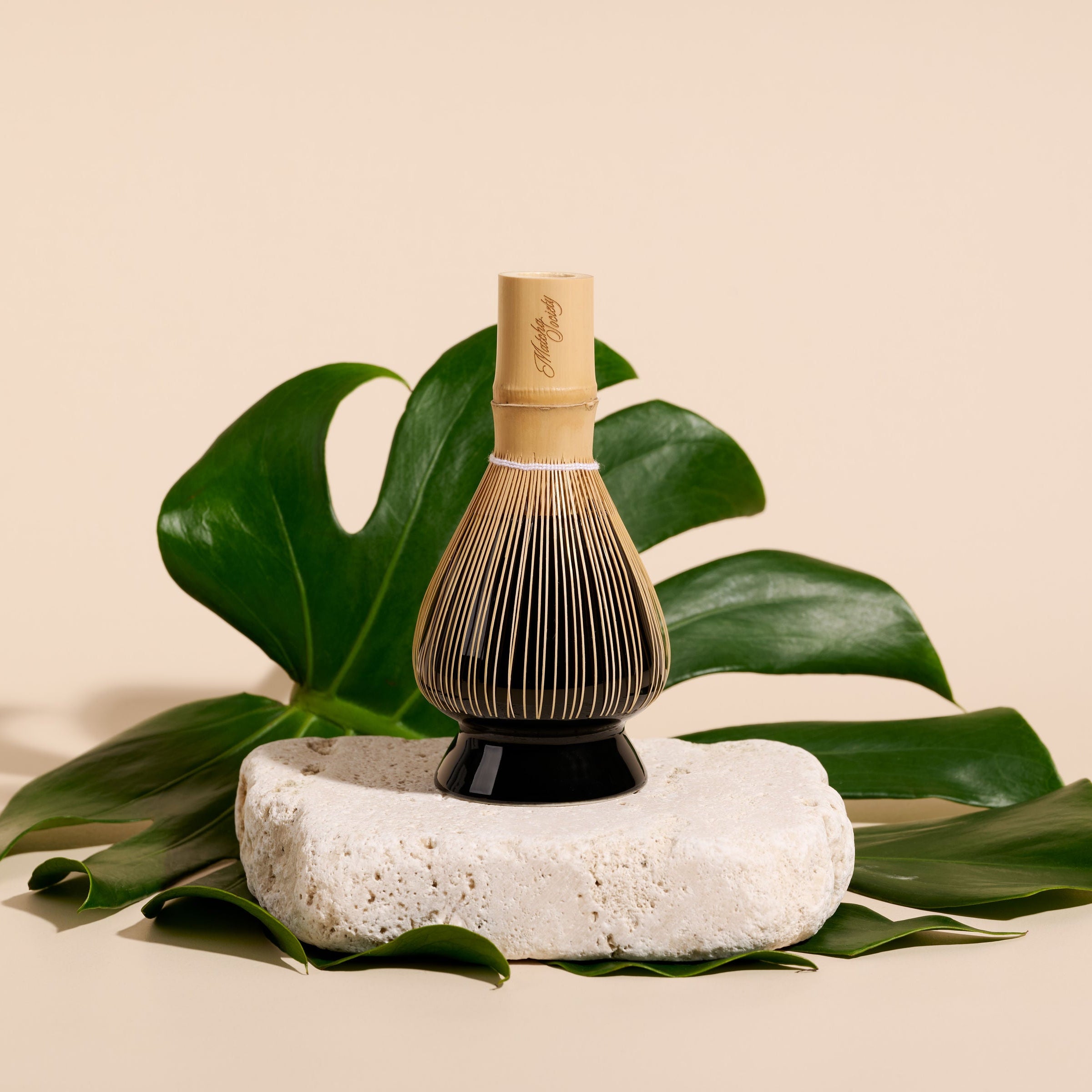 A bamboo whisk with a black stand, displayed on a white stone pedestal with green monstera leaves against a beige background.