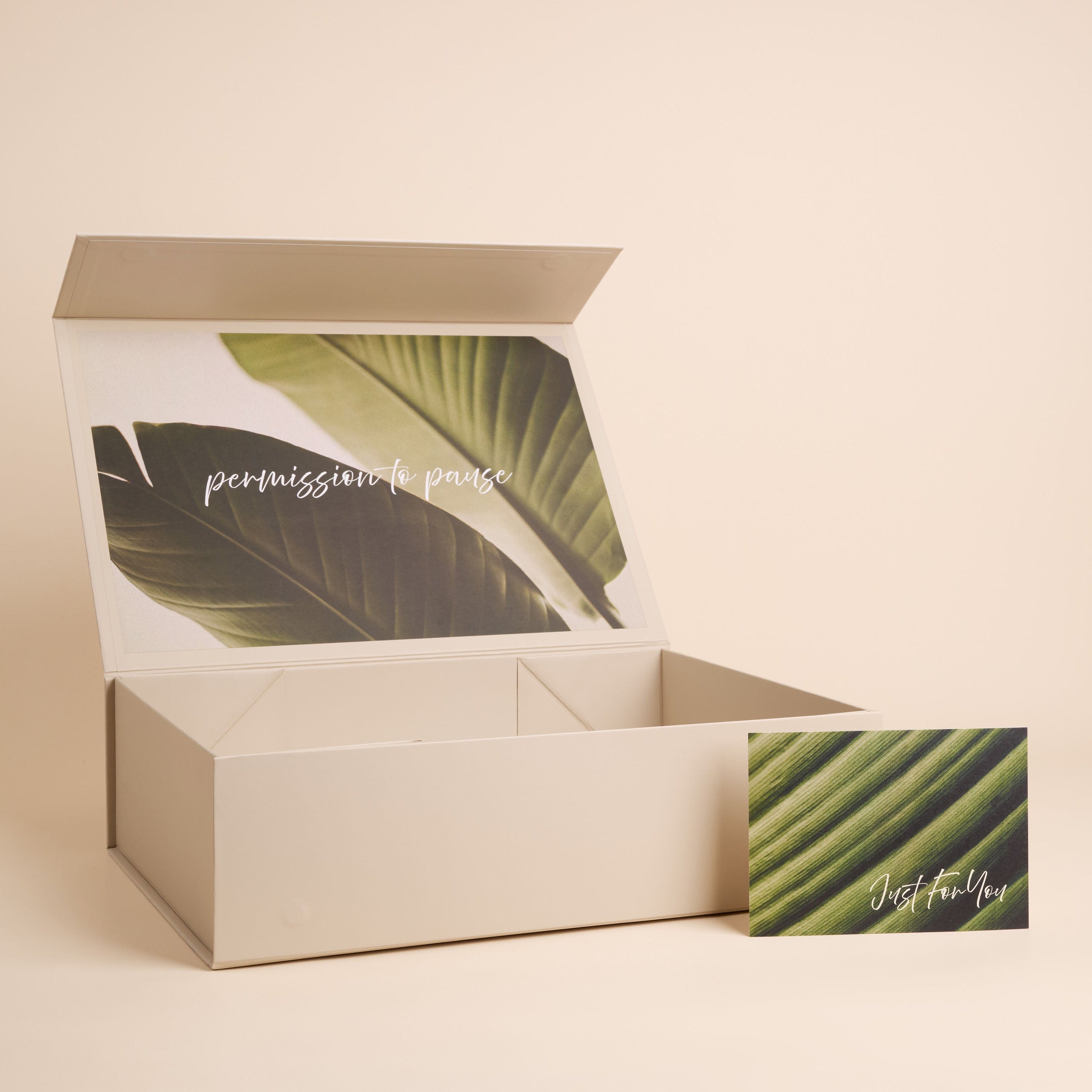 A luxury PAVILION gift box with its lid open, printed with a large green tropical leaf and the words 'permission to pause.' A matching small rectangular card with the same leaf print and Just For You message rests beside the box on a warm beige background.