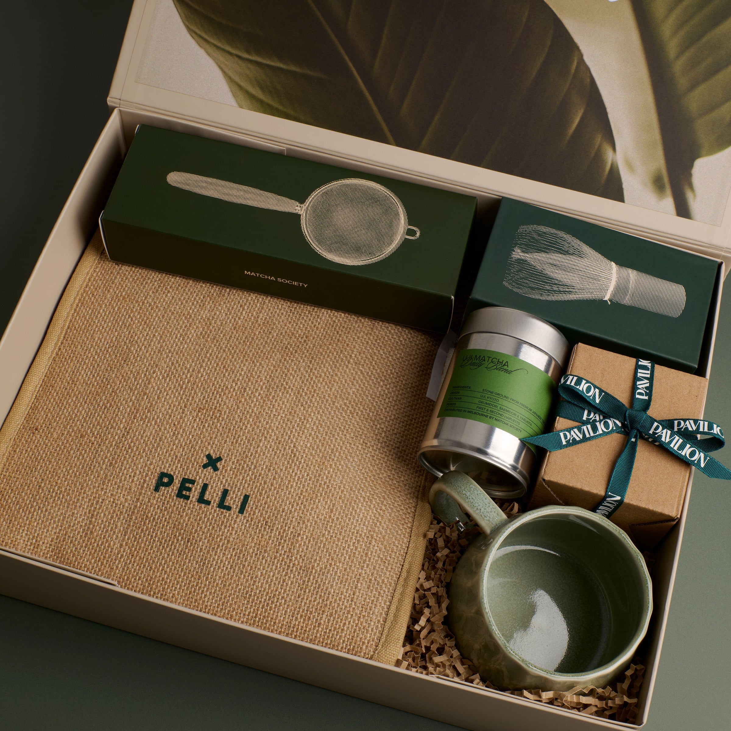 An open matcha gift box set on a dark green background, containing a beige jute Pelli insulated tote, a green matcha canister from Matcha Society, a ceramic cup, and a strainer in its packaging.