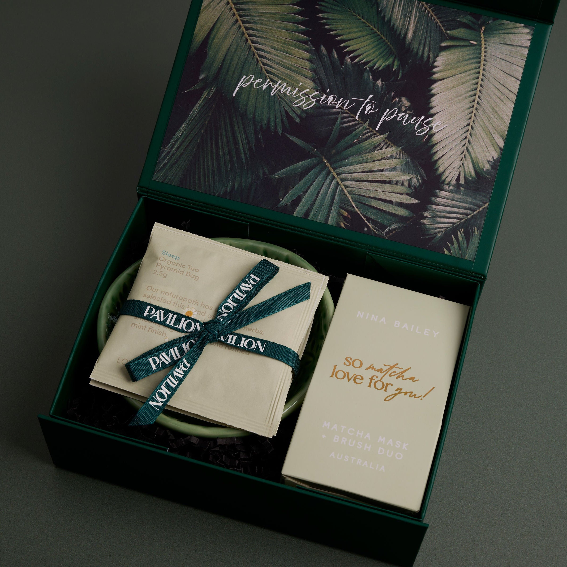 An open dark green gift box with a tropical palm leaf pattern on the inside lid, containing a trio of beauty teas, a ceramic dish and a matcha clay face mask set with mask brush. 